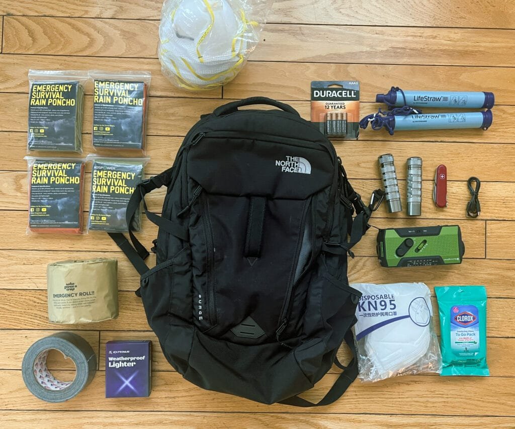 A black North Face backpack is surrounded by emergency preparedness items including fire blankets, smoke-filtering masks, high-intensity flashlights, a thermal blanket, a Swiss Army knife, a portable radio, KN95 masks, disinfectant wipes, duct tape, a weatherproof lighter, batteries, and other survival tools, all arranged on a wooden surface.